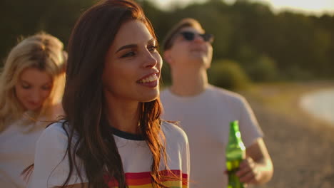 A-young-happy-woman-with-long-dark-hair-in-a-short-white-t-shirt-is-dancing-on-the-open-air-party-with-beer.-She-smiles-and-touchs-her-hair-and-enjoys-party-time-on-the-lake-coast-at-sunrise.